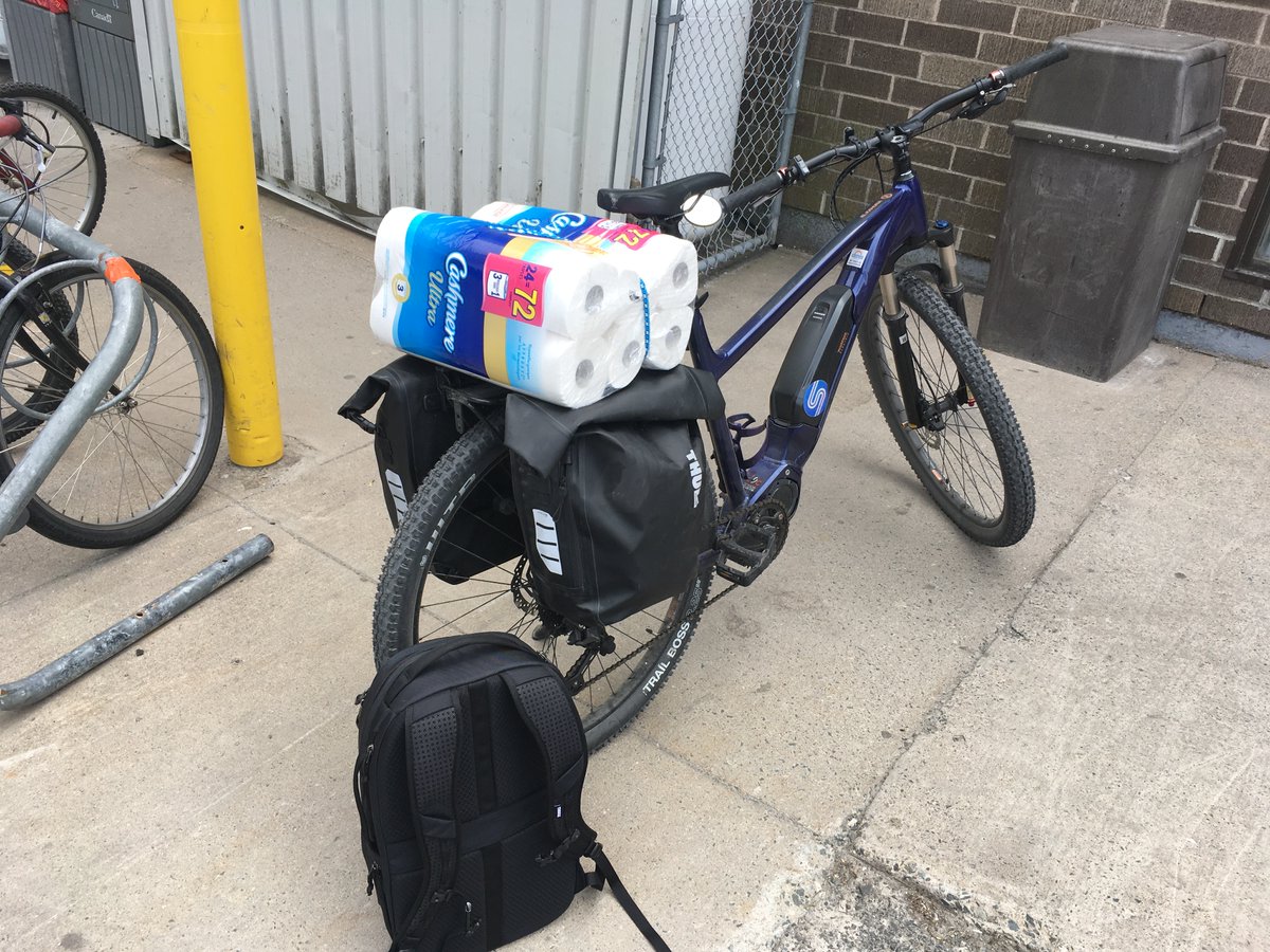 Ebike parked near a grocery store has full pannier bags and a big package of toilet paper bungie-corded to the rack over the back tire.